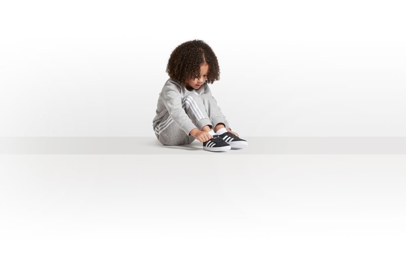 A young person with curly hair sits on the floor, wearing a gray sweatshirt and black sneakers, against a plain white background.