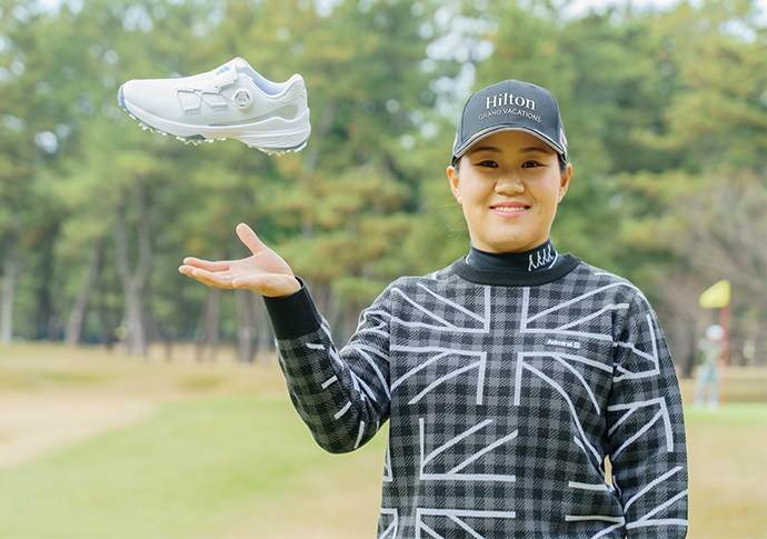 A smiling person wearing a Hilton cap and a checkered jacket is holding up a white sneaker in a grassy outdoor setting with trees in the background.