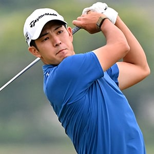 A golfer in a white cap and blue shirt is swinging a golf club on a grassy golf course with trees in the background.
