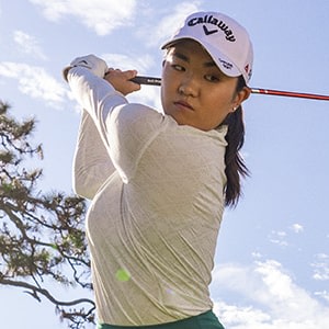 A young woman in a white golf shirt and cap is swinging a golf club on a sunny day with trees and a cloudy sky in the background.