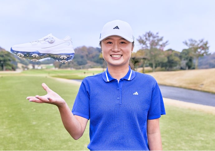 A smiling person in a blue polo shirt and white cap stands on a golf course, holding up a golf shoe in the foreground.