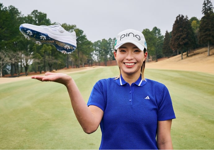 A smiling woman in a blue golf polo and a white golf cap stands on a lush green golf course surrounded by trees.