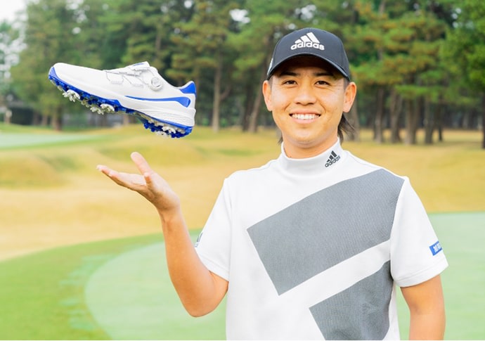 A smiling man in an Adidas cap and athletic clothing holds up a golf shoe on a grassy golf course surrounded by trees.
