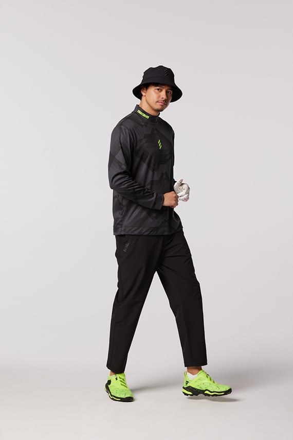 A person wearing a black hat, black hoodie, and black pants stands in a plain white background, holding a gaming controller in their hands.