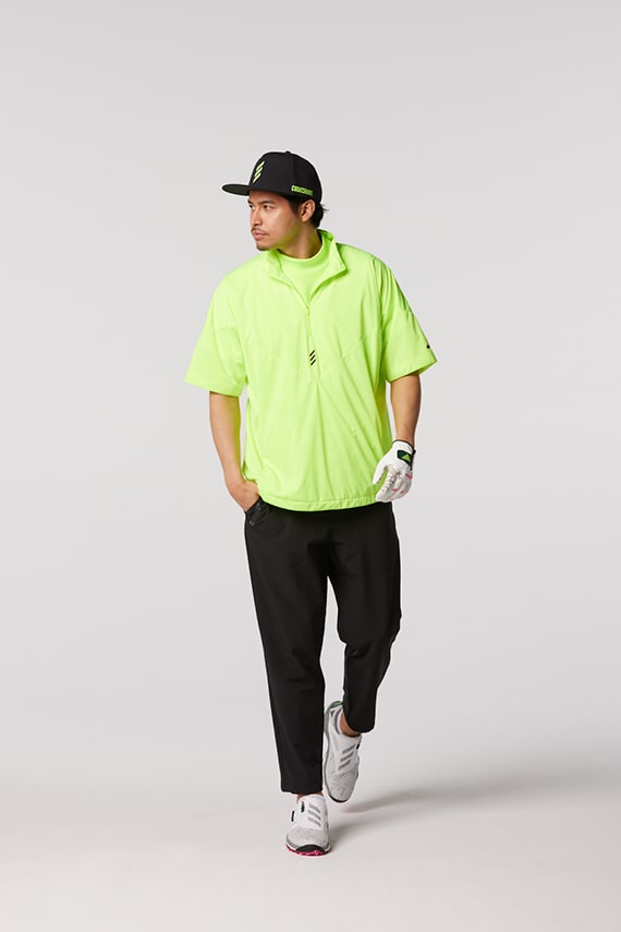 A person wearing a bright neon green shirt, black pants, and a black cap is walking against a plain white background.