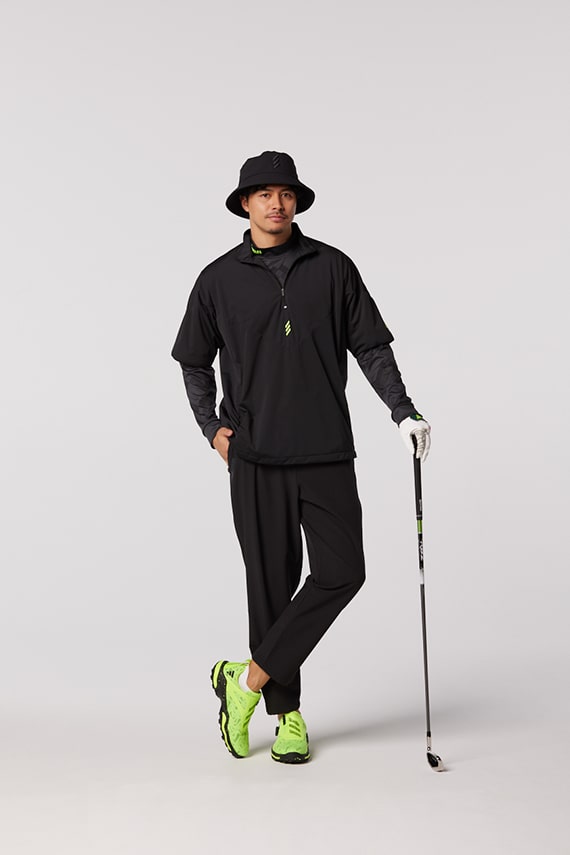 A person wearing a black outfit, including a hat, stands holding a walking stick and wearing bright green sneakers against a plain white background.