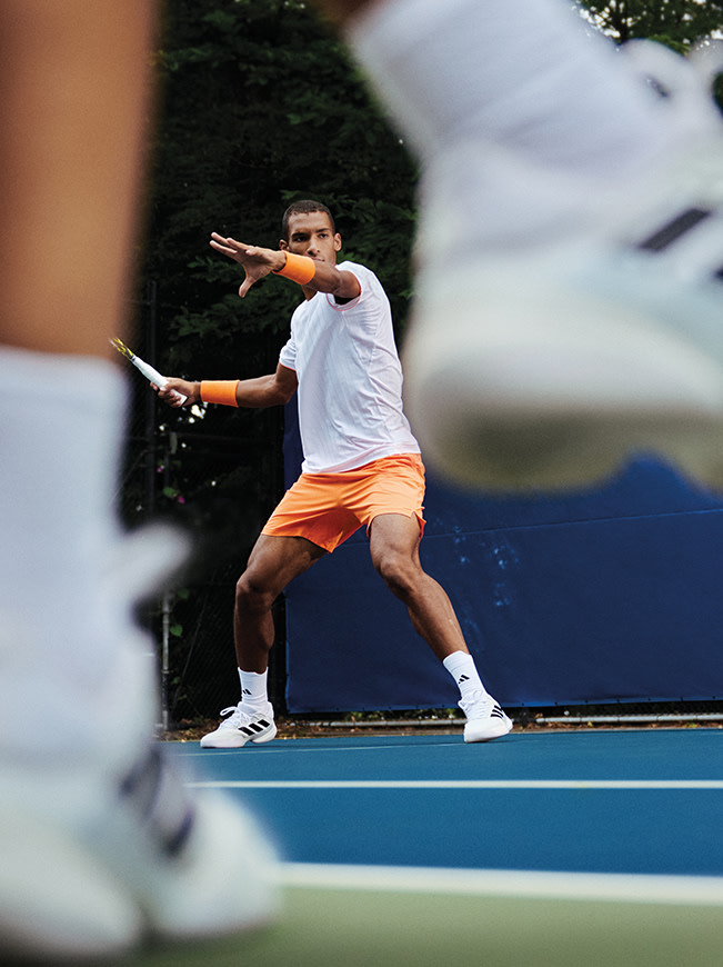 A person in white and orange sportswear is playing tennis on a blue tennis court surrounded by trees.