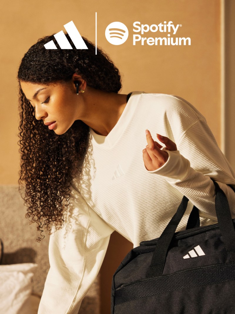 A young woman with curly hair wearing a white sweater and holding a black Adidas bag, with the Spotify Premium logo visible in the background.