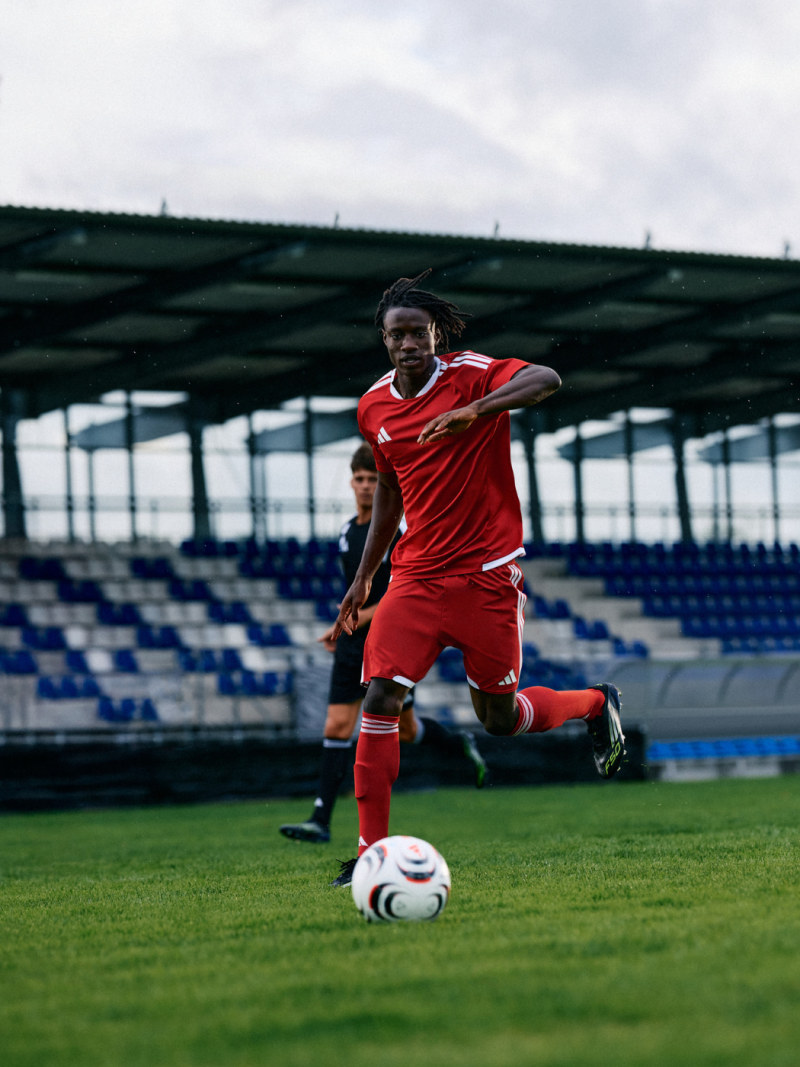 A soccer player in a red uniform is running with the ball on a grassy field, with a stadium and bleachers visible in the background.