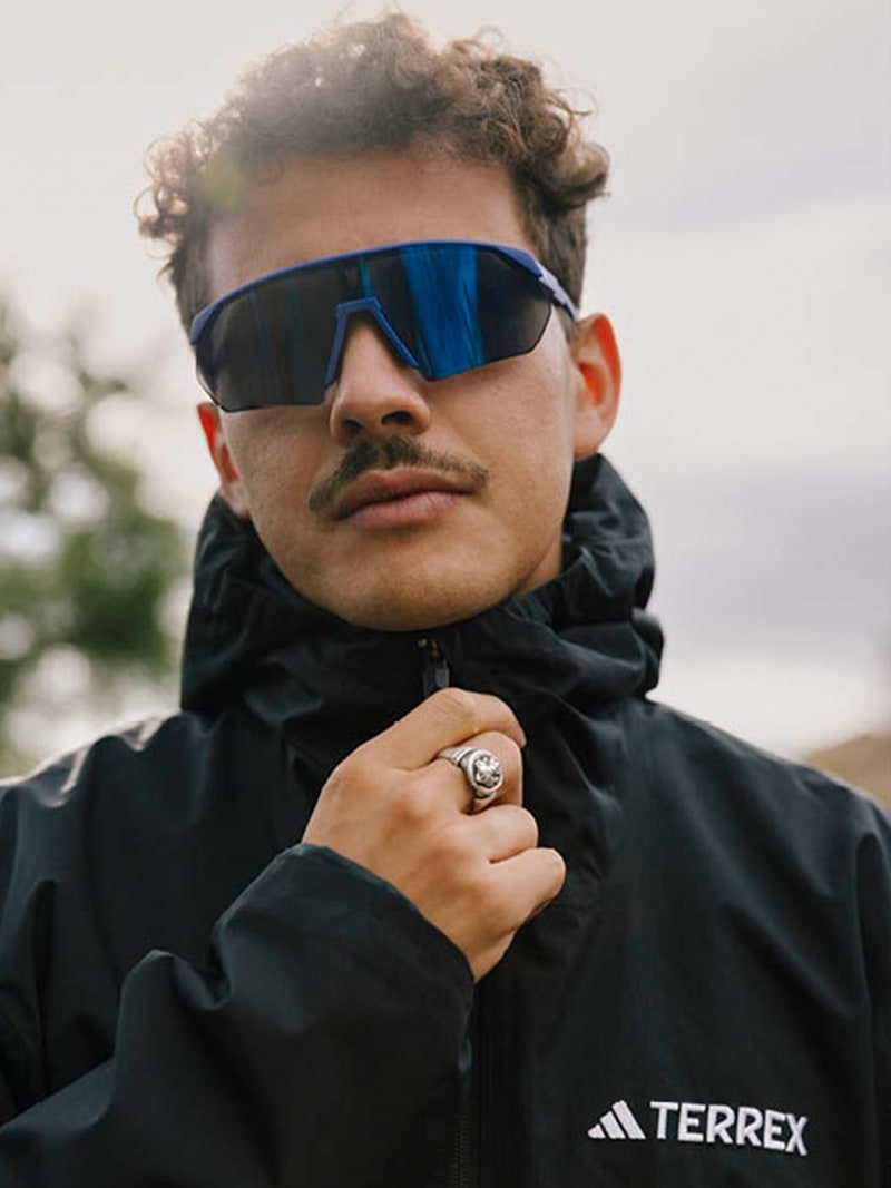 A person wearing a black jacket and sunglasses stands in an outdoor setting with a cloudy sky in the background.