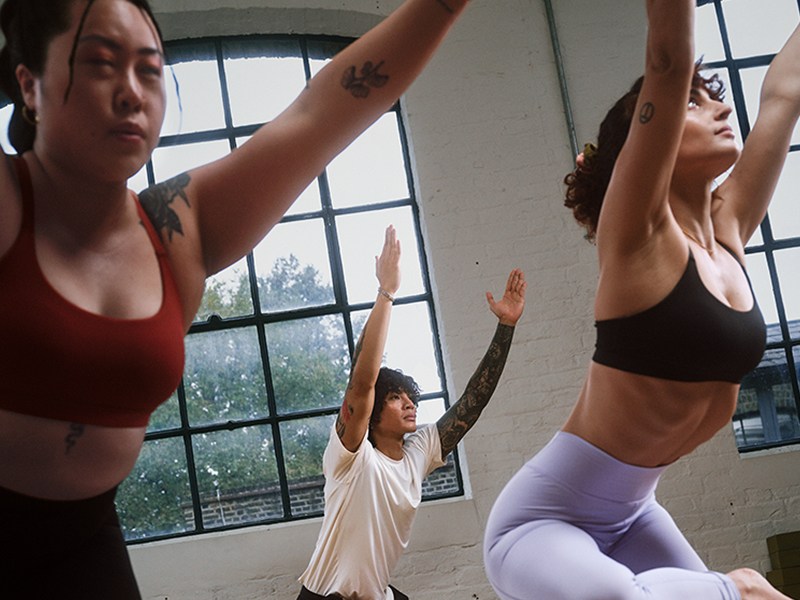 The image shows three women practicing yoga poses in a studio setting with large windows overlooking a scenic outdoor view.