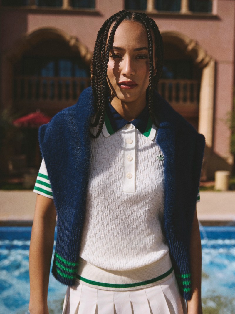 The image shows a person with braided hair wearing a white shirt, a blue sweater, and standing in front of a building.