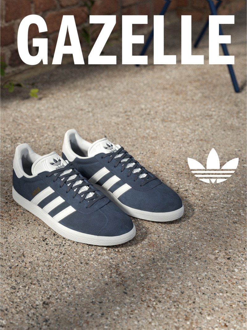 A pair of navy blue and white Adidas Gazelle sneakers resting on a gravel surface, with a blurred background of greenery.
