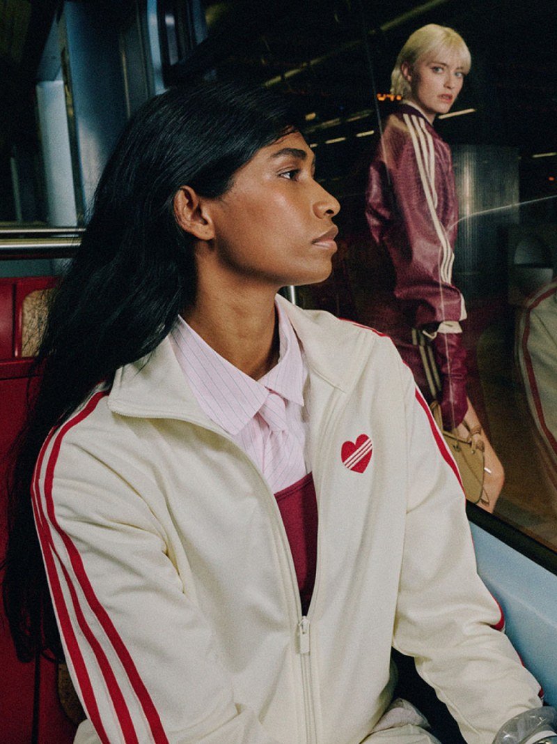The image shows a young woman with dark hair wearing a white jacket with red stripes, sitting in what appears to be a train or subway car, with another person visible in the background.