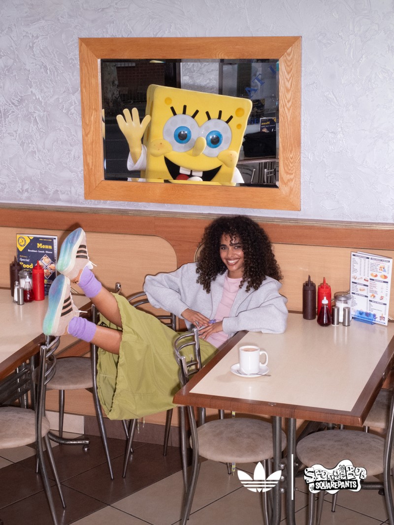 A young woman with curly hair sits at a table in a room with a framed image of the cartoon character SpongeBob SquarePants on the wall above.