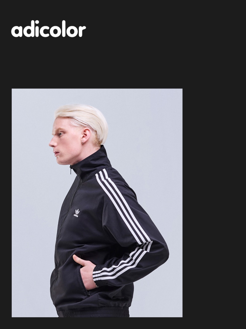 A person wearing a black Adidas jacket with white stripes stands against a plain gray background.