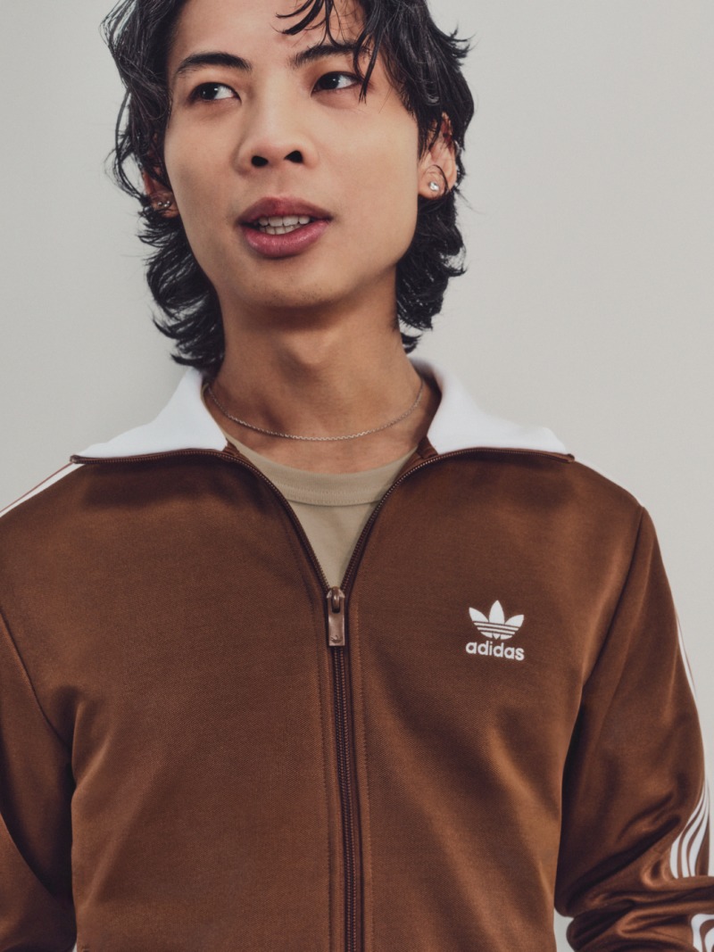 A person crouches wearing a brown Beckenbauer tracksuit with white stripes and looks at the camera.