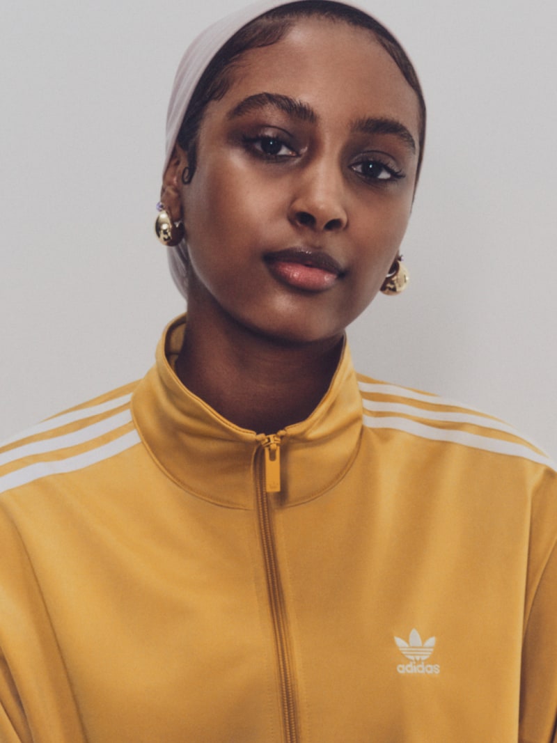 A person stands in a light brown Firebird track top with white stripes, facing the camera.