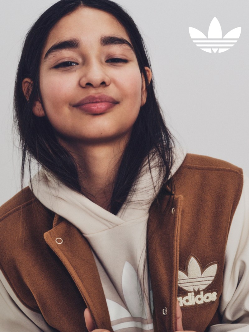 Person with long dark hair wearing a brown and cream Adidas hoodie, head tilted.