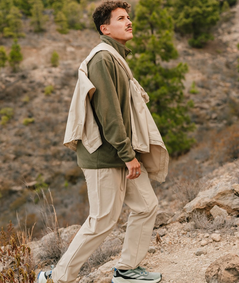 A person wearing a beige and olive-colored outfit stands on a rocky, mountainous terrain with lush vegetation in the background.