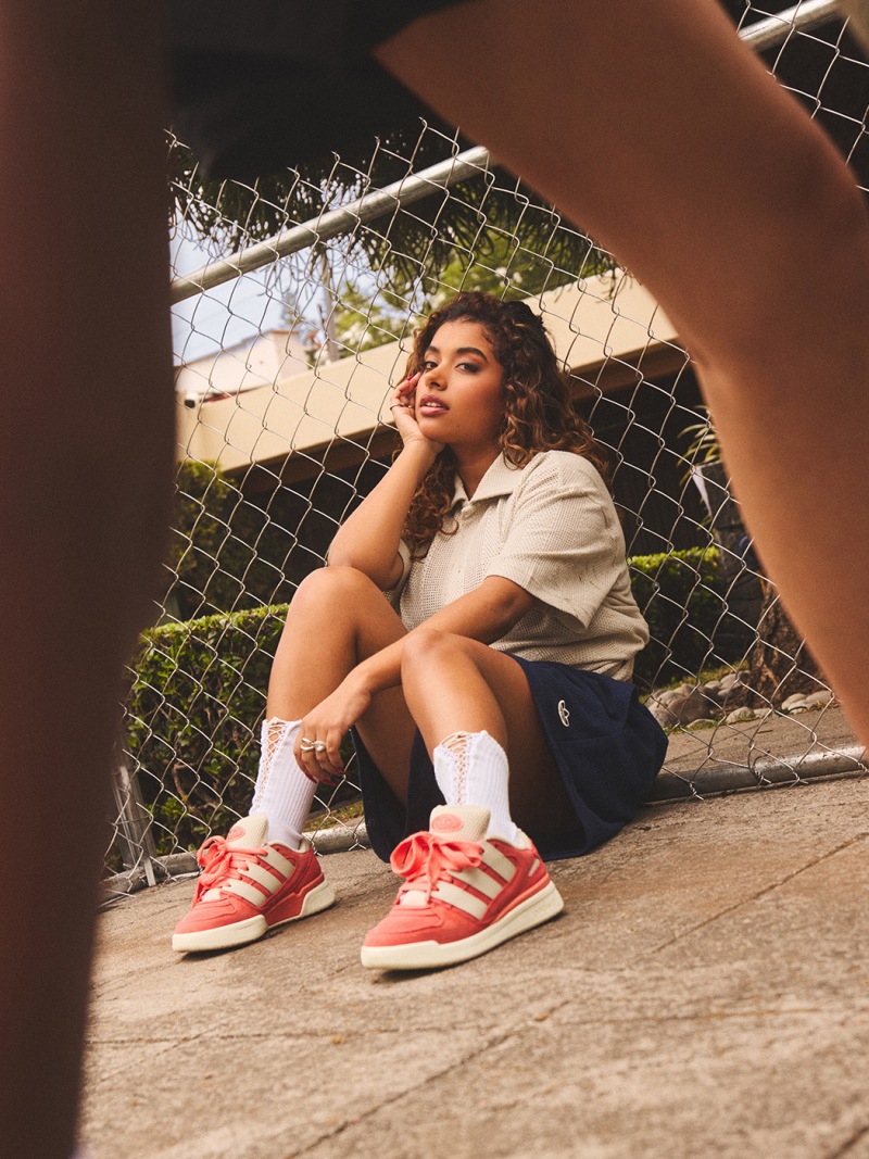 A person with curly hair sits on the ground, wearing a light-colored shirt and red sneakers, against a backdrop of a wire fence and greenery.