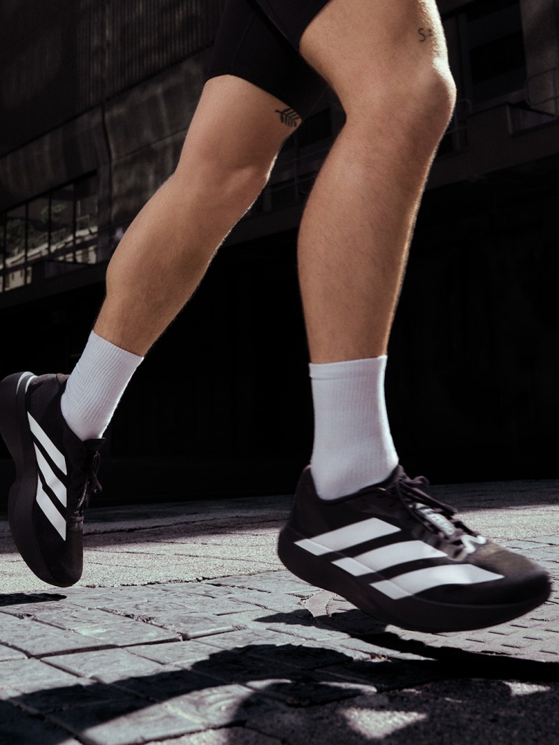 The image shows the lower body of a person wearing black and white athletic shoes and white socks, standing on a dark, textured surface.
