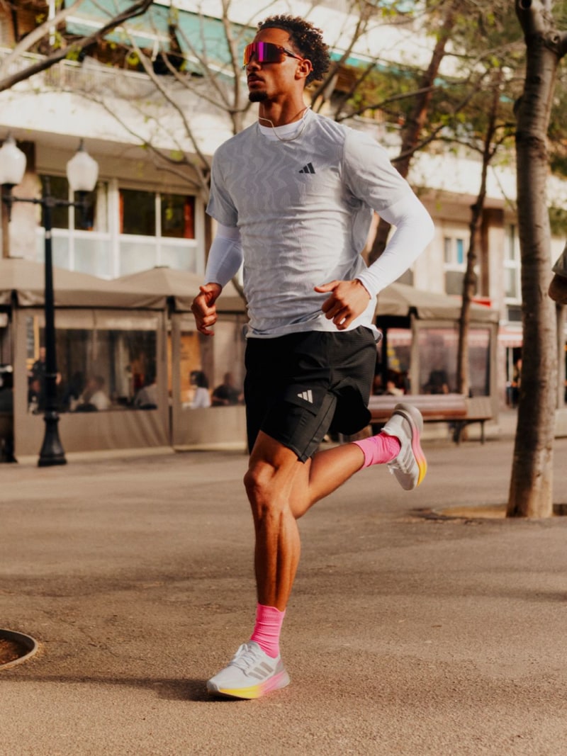 A person wearing athletic clothing and sunglasses is running on a paved path in an urban setting, with buildings and trees visible in the background.