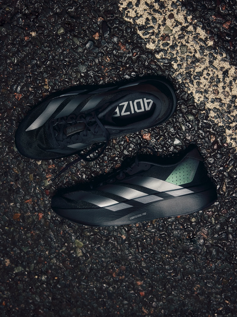 A pair of black and gray athletic shoes rests on a gravel-covered surface, with the brand name "Adidas" prominently displayed on the side of the shoes.