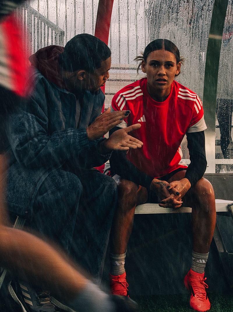 The image shows two individuals, one wearing a red jersey and the other in a dark jacket, sitting together on a bench in what appears to be a locker room or similar indoor setting.