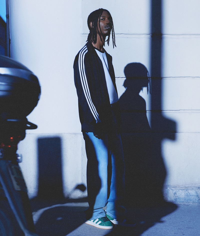 A person with dreadlocks wearing a black jacket with white stripes stands in a dimly lit setting, with the silhouette of another person visible in the background.