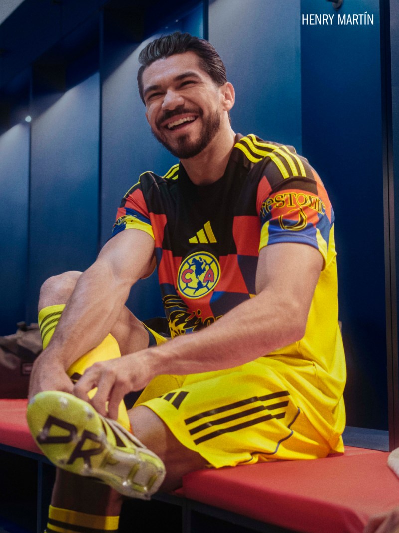 A smiling man wearing a yellow and black soccer jersey sits on a bench in what appears to be a locker room or training facility.