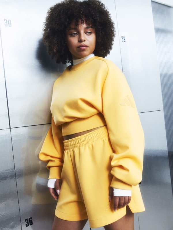 Uma jovem com cabelo escuro encaracolado usando uma camisola e calções amarelos brilhantes destaca-se em frente a um cacifo cinza.