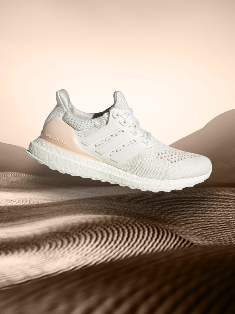 A white and beige athletic shoe rests on a desert landscape with undulating sand dunes in the background.
