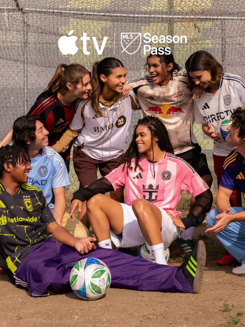 A group of diverse female soccer players, some wearing jerseys of different teams, sitting together on the field and interacting with each other.