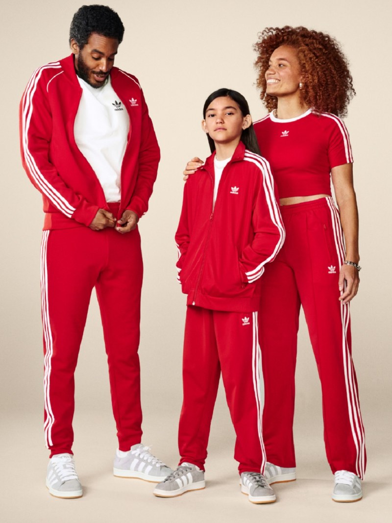 A family of three, a man, a woman, and a young girl, all wearing matching red and white Adidas tracksuits, posing together against a plain background.