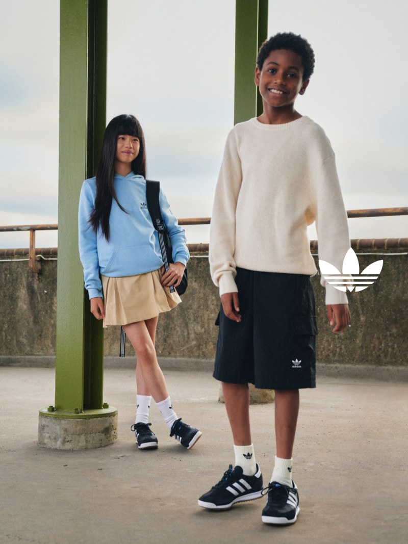Two young people, a girl and a boy, standing together outdoors against a backdrop of a concrete structure and a cloudy sky.