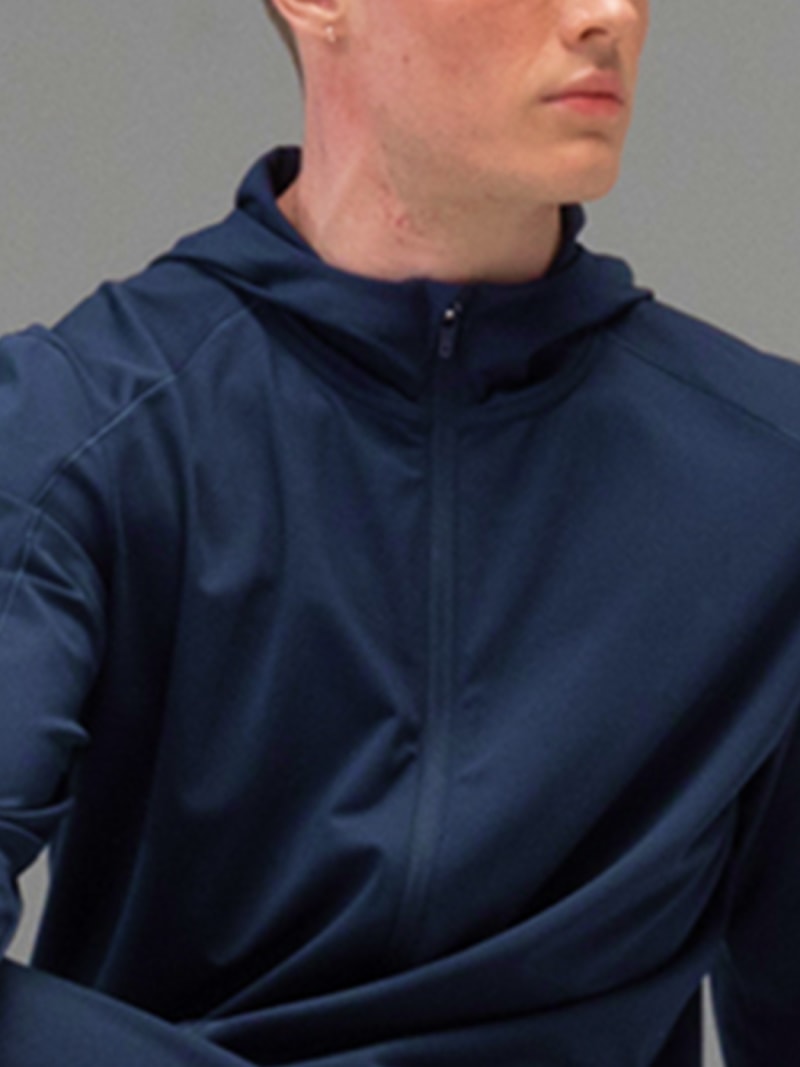 A person wearing a navy blue hooded jacket stands against a plain gray background.