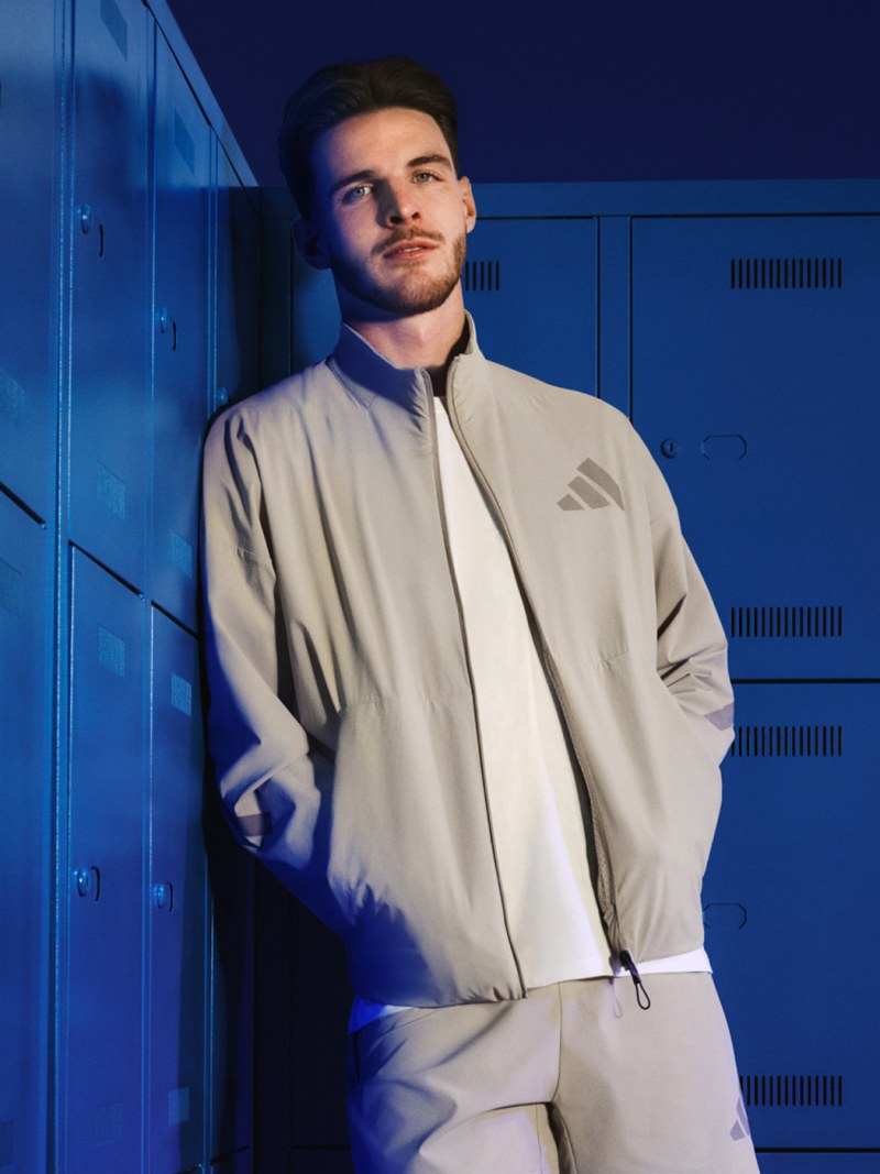A young man wearing a beige jacket stands in front of blue lockers, his expression thoughtful and his posture relaxed.