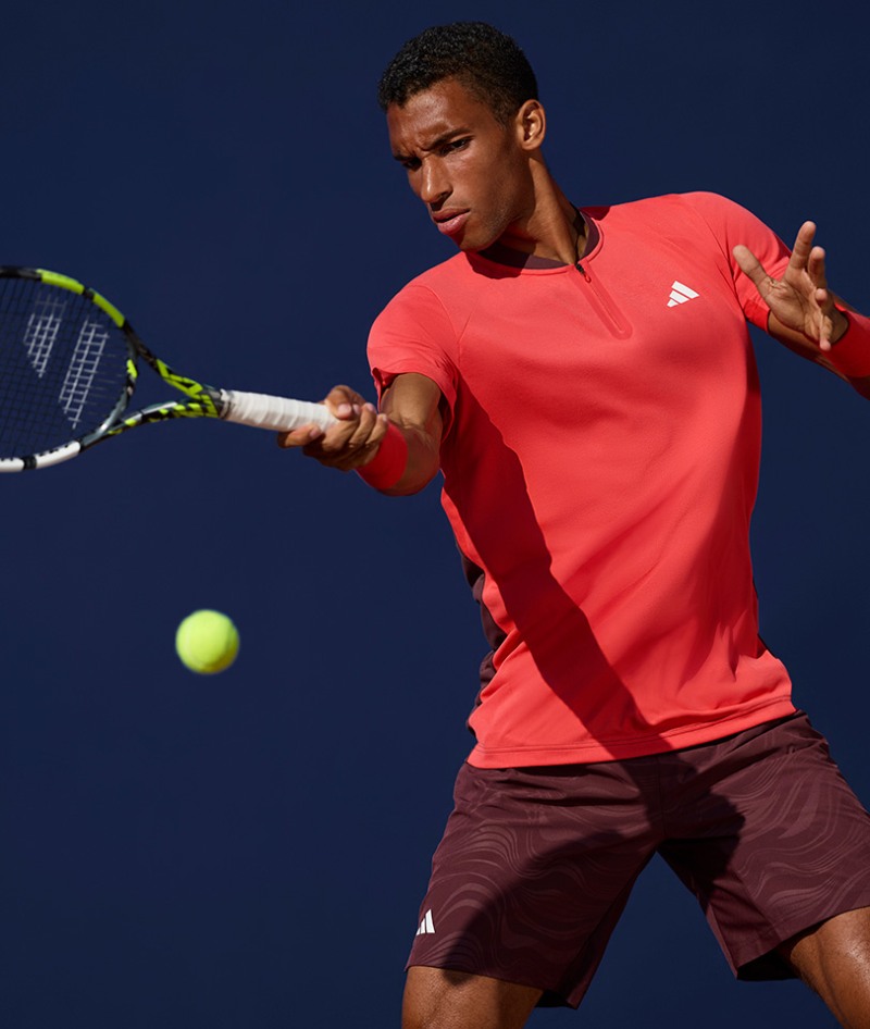 A tennis player in a red shirt and brown shorts is swinging a tennis racket while a tennis ball is in motion in the foreground, with a dark blue background.