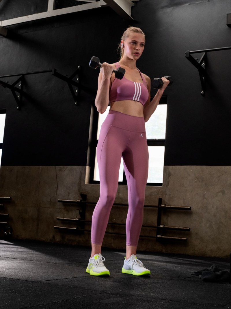 A woman in a pink sports bra and leggings stands in a dimly lit gym, holding weights and facing the camera.