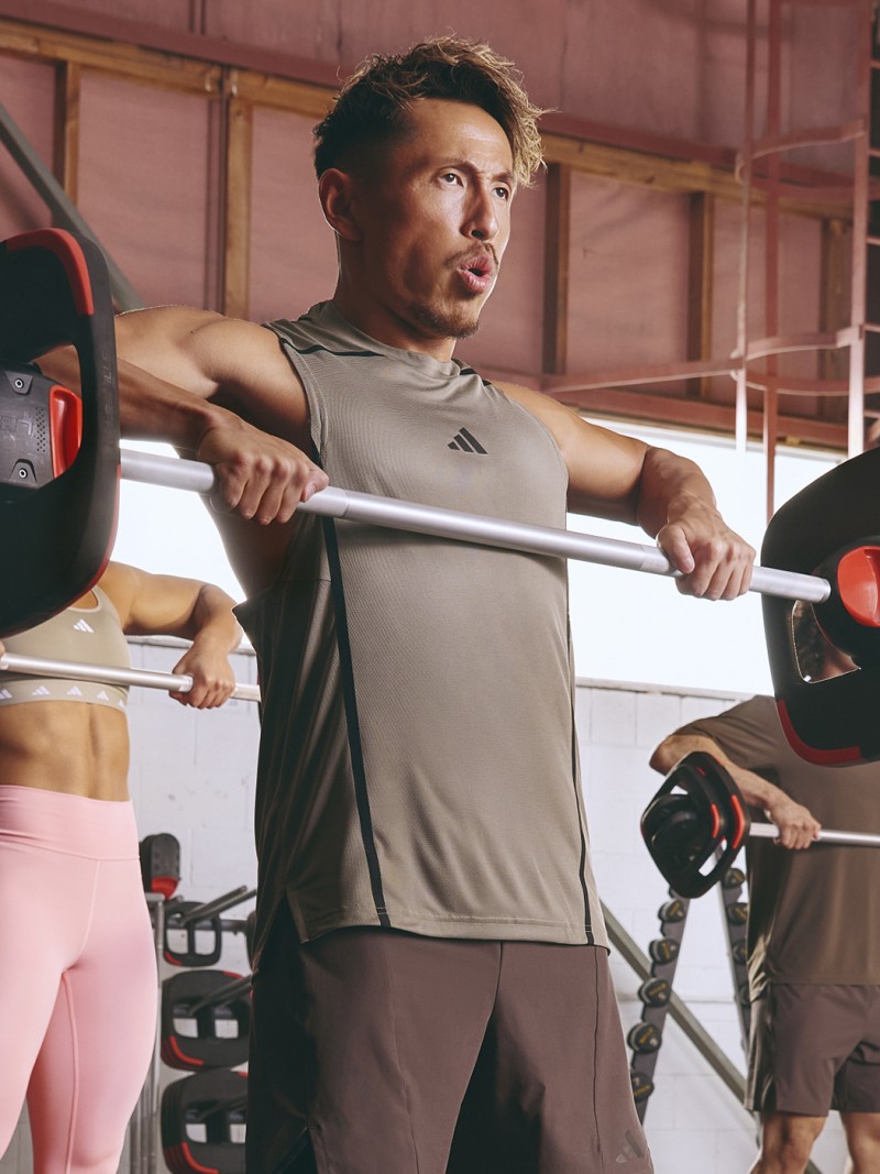 A man in a gray athletic shirt stands in a gym, surrounded by exercise equipment and weights.
