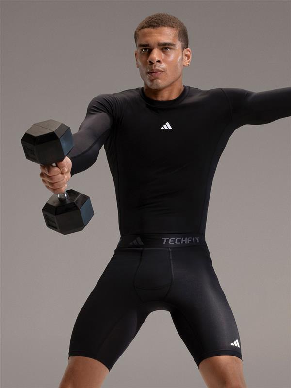 A man in a black athletic outfit is holding two black dumbbells and standing against a plain gray background.