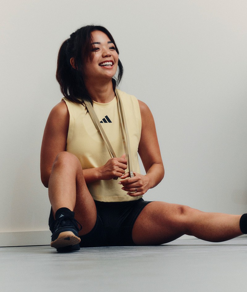 A smiling young woman in a yellow top and black shorts sits on a white floor, her hair falling over her shoulders.
