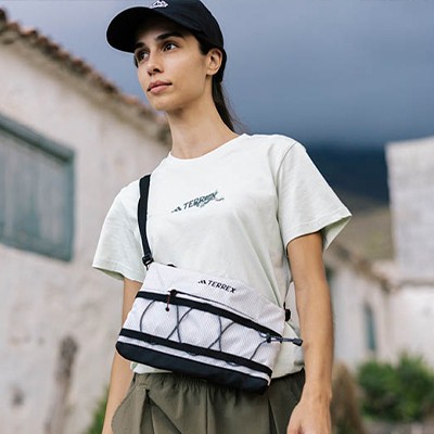 A young woman wearing a white t-shirt and a black baseball cap stands in front of a building, with a scenic background of a body of water and a cloudy sky.