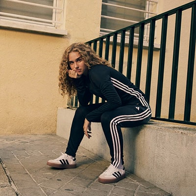 A person with curly hair wearing a black tracksuit and Adidas sneakers is sitting on a ledge against a building with windows.
