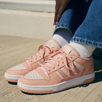 The image shows a pair of pink and white sneakers on a wooden floor, with the person's legs and jeans visible in the foreground.