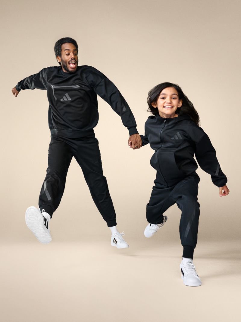 Two people, a man and a woman, are jumping and smiling while wearing matching black athletic outfits and white sneakers against a plain beige background.