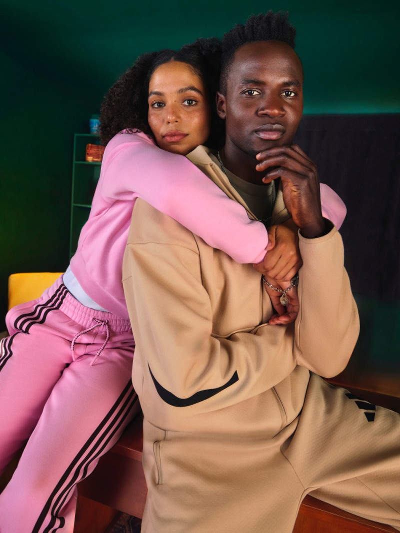 The image shows a couple embracing, with the woman wearing a pink outfit and the man wearing a beige coat. They appear to be in a cozy, dimly lit setting with a green background.