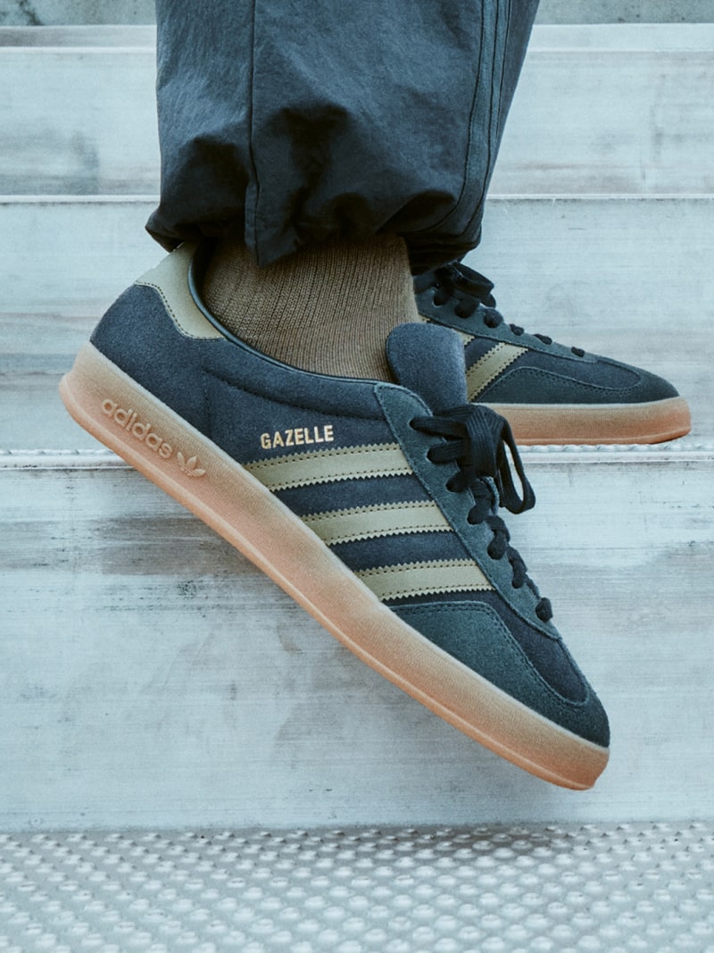 A pair of gray and beige Adidas Gazelle sneakers with a gum sole, worn by someone standing on a tiled floor.