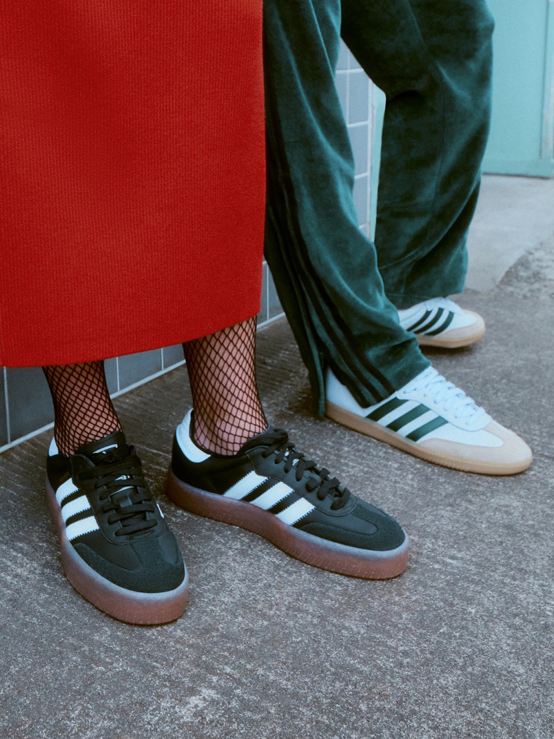 The image shows a person wearing dark green pants, fishnet stockings, and a pair of black and white sneakers with a gum sole standing in front of a bright red background.